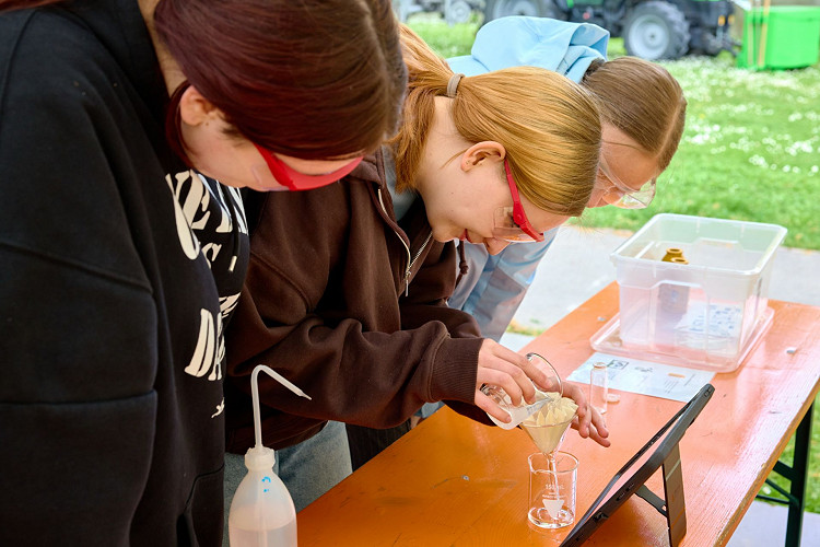 Drei Jugendliche führen bei einem Workshop von Explore Science im Freien ein naturwissenschaftliches Experiment durch. Mit Schutzbrillen ausgestattet arbeiten sie konzentriert an einem Versuchsaufbau zur Untersuchung einer Flüssigkeit. Neben ihnen stehen verschiedene Materialien sowie ein Tablet zur Begleitung des Experiments.