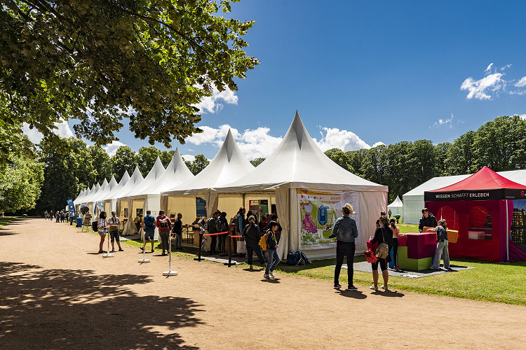 Mehrere weiße Mitmachzelte und ein roter Pavillon auf dem Gelände von Explore Science im Herzogenriedpark Mannheim. Besucher:innen schlendern entlang der Zelte unter schattigen Bäumen. Der Himmel ist blau mit vereinzelten Wolken.