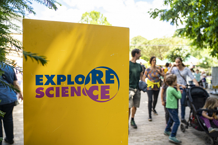 gelbes Schild mit pink blauem Logo von Explore Science mit Besucherinnen und Besuchern im Hintergrund